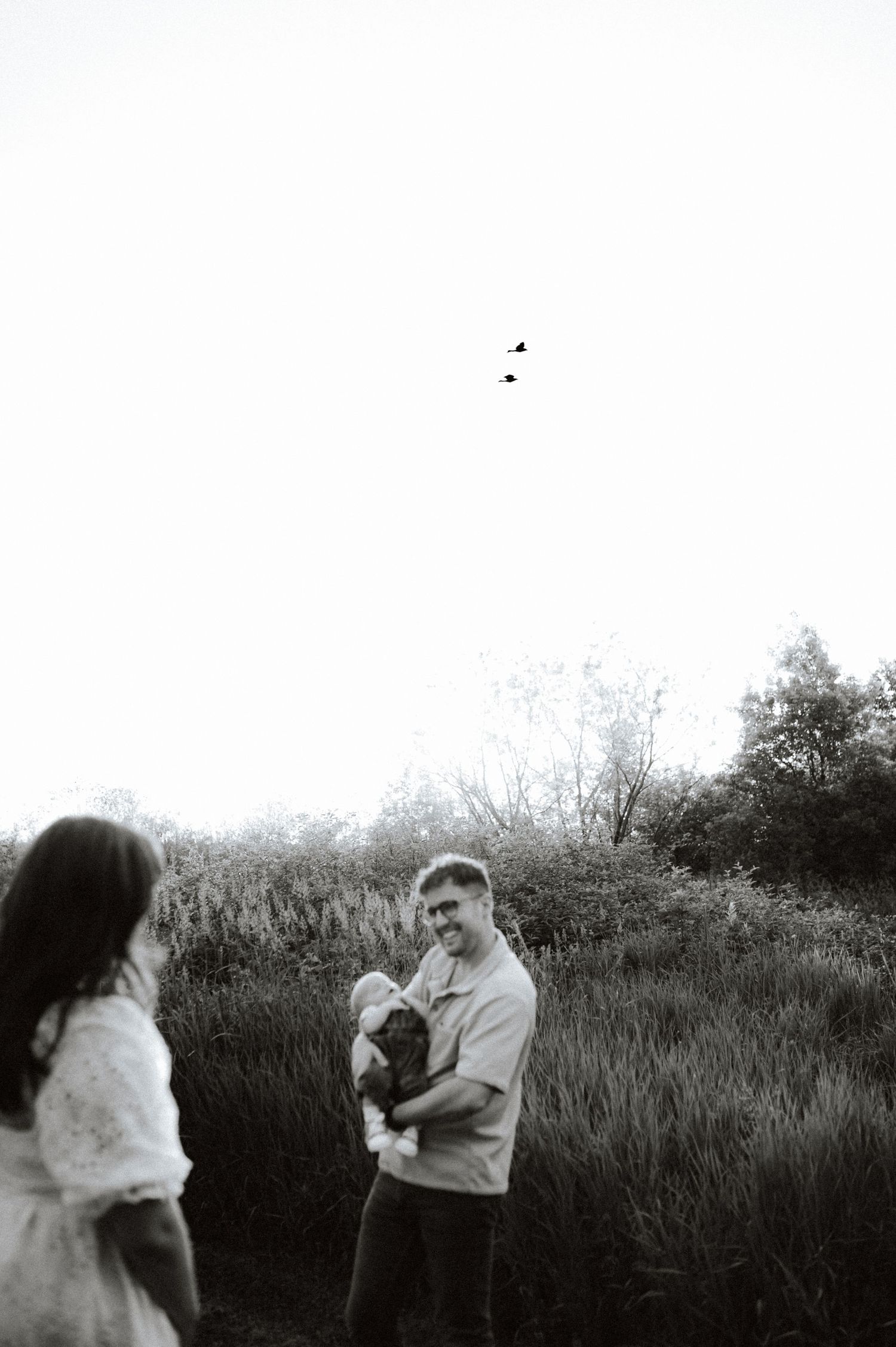 A black and white outdoor photo showing people talking and holding a small dog in a grassy natural setting.
