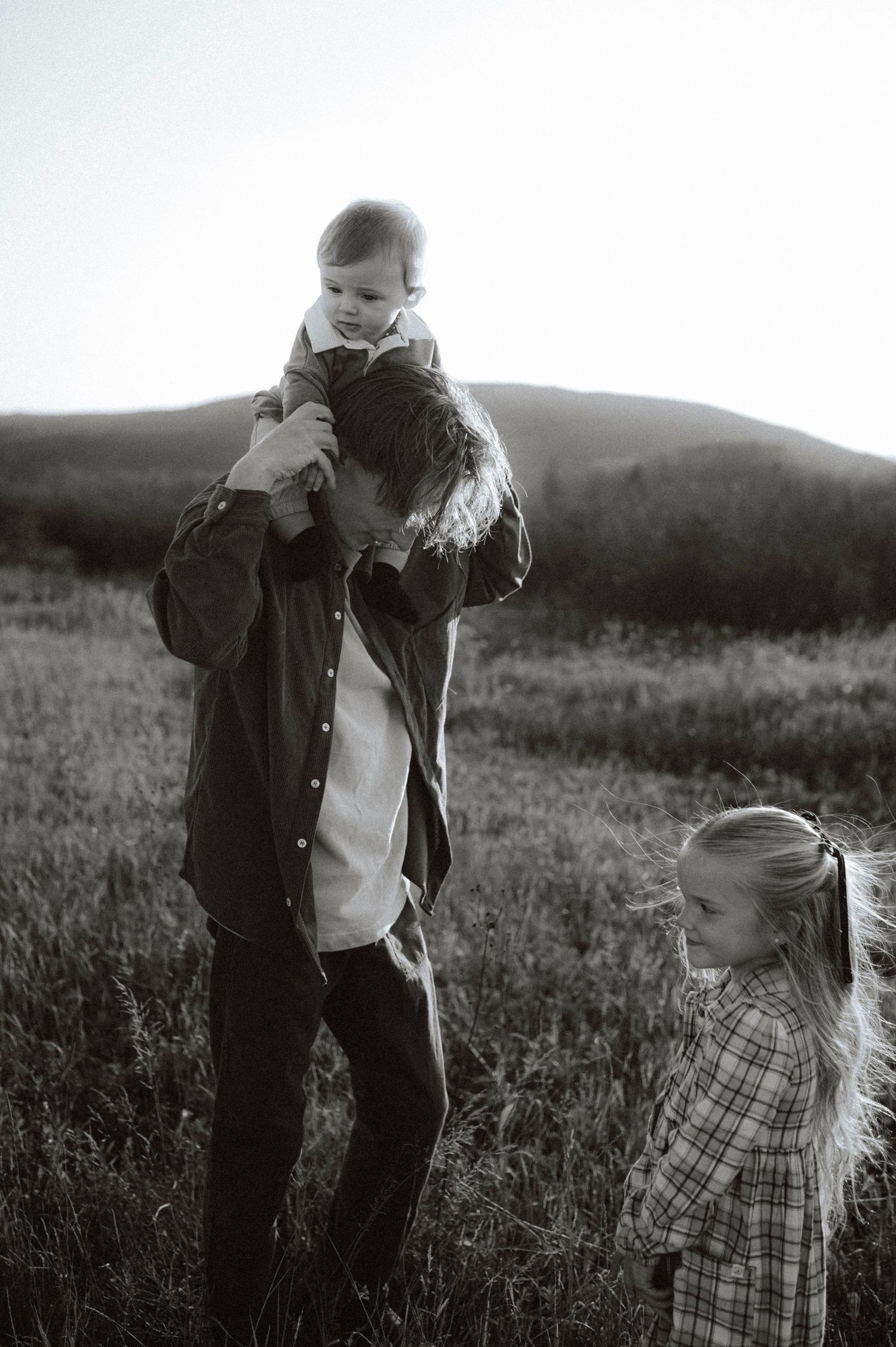 Black and white photo of a heartwarming family moment in a rural field with mountains in the background.