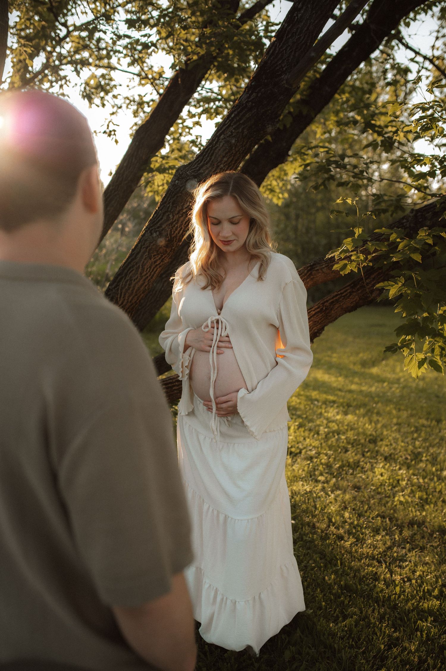 A tender maternity photoshoot at golden hour captures an intimate outdoor moment under a large tree.