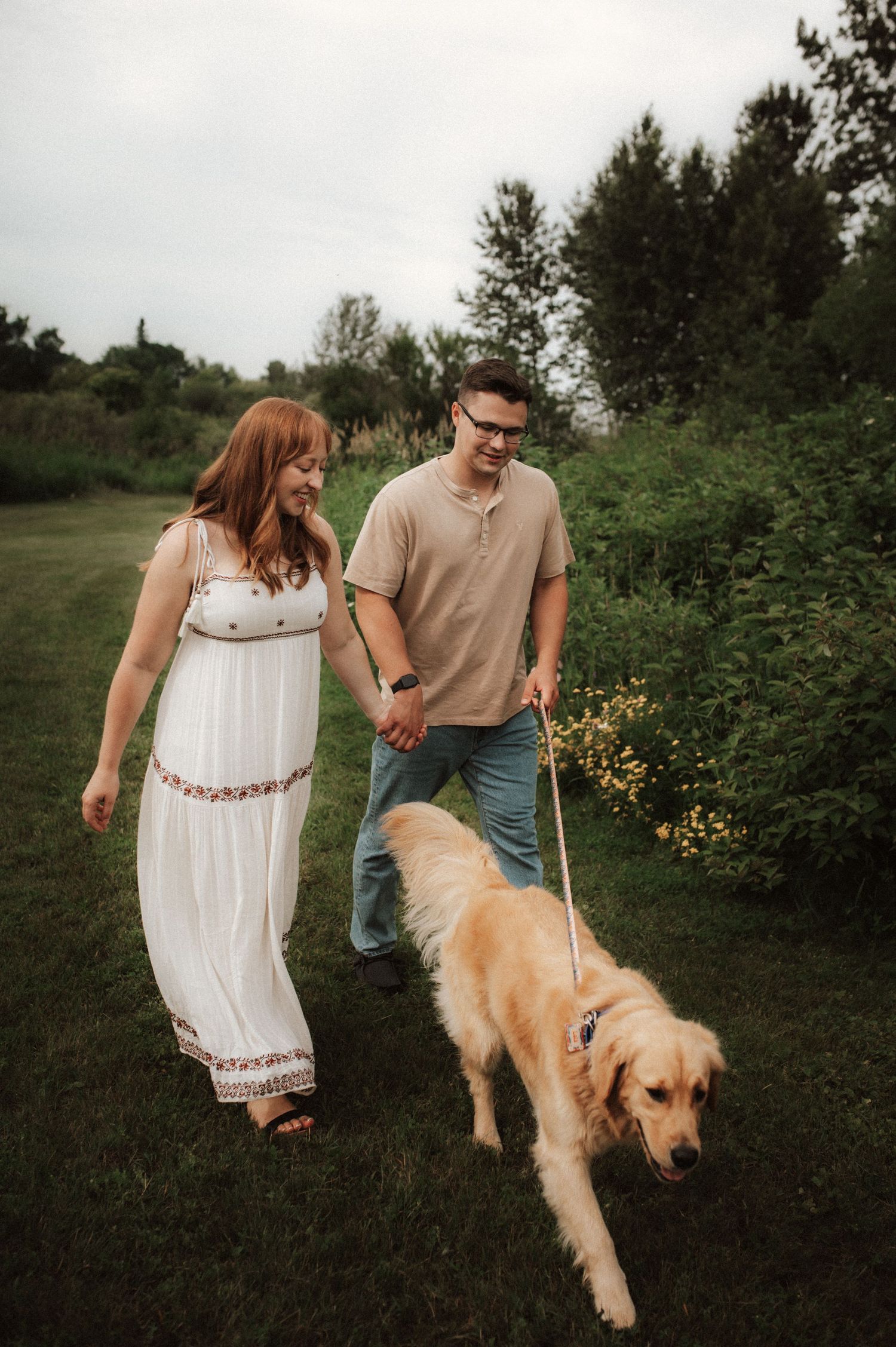A couple walks through a green field with their golden retriever enjoying a peaceful outdoor moment together.