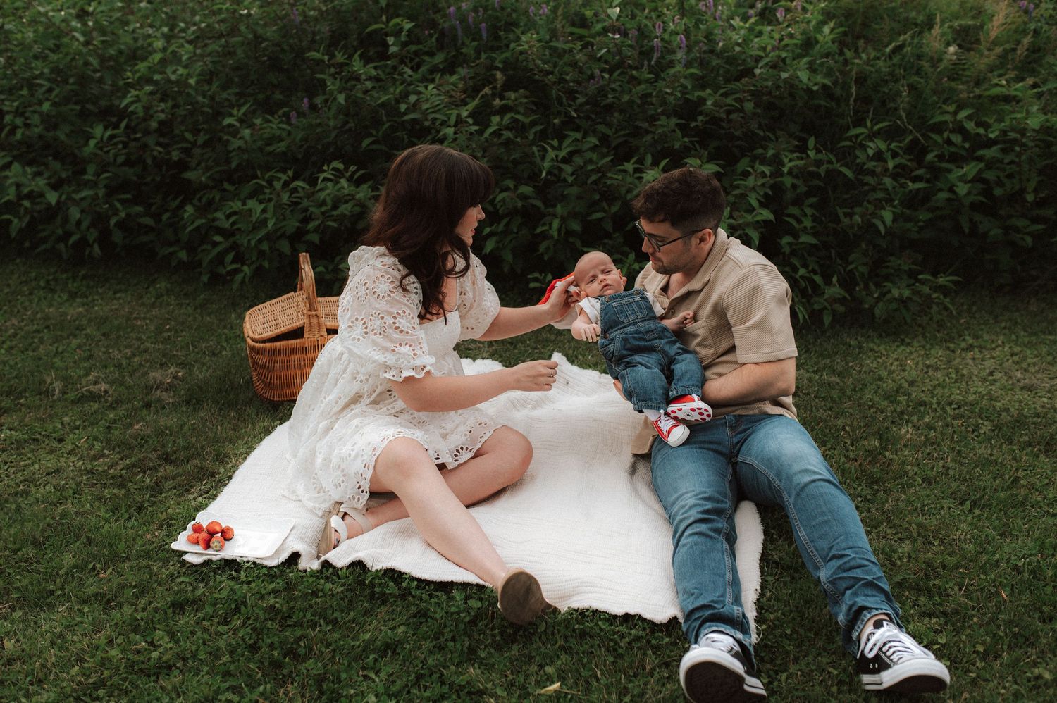 A family sits together on a white blanket in a grassy outdoor area while engaged in various activities.