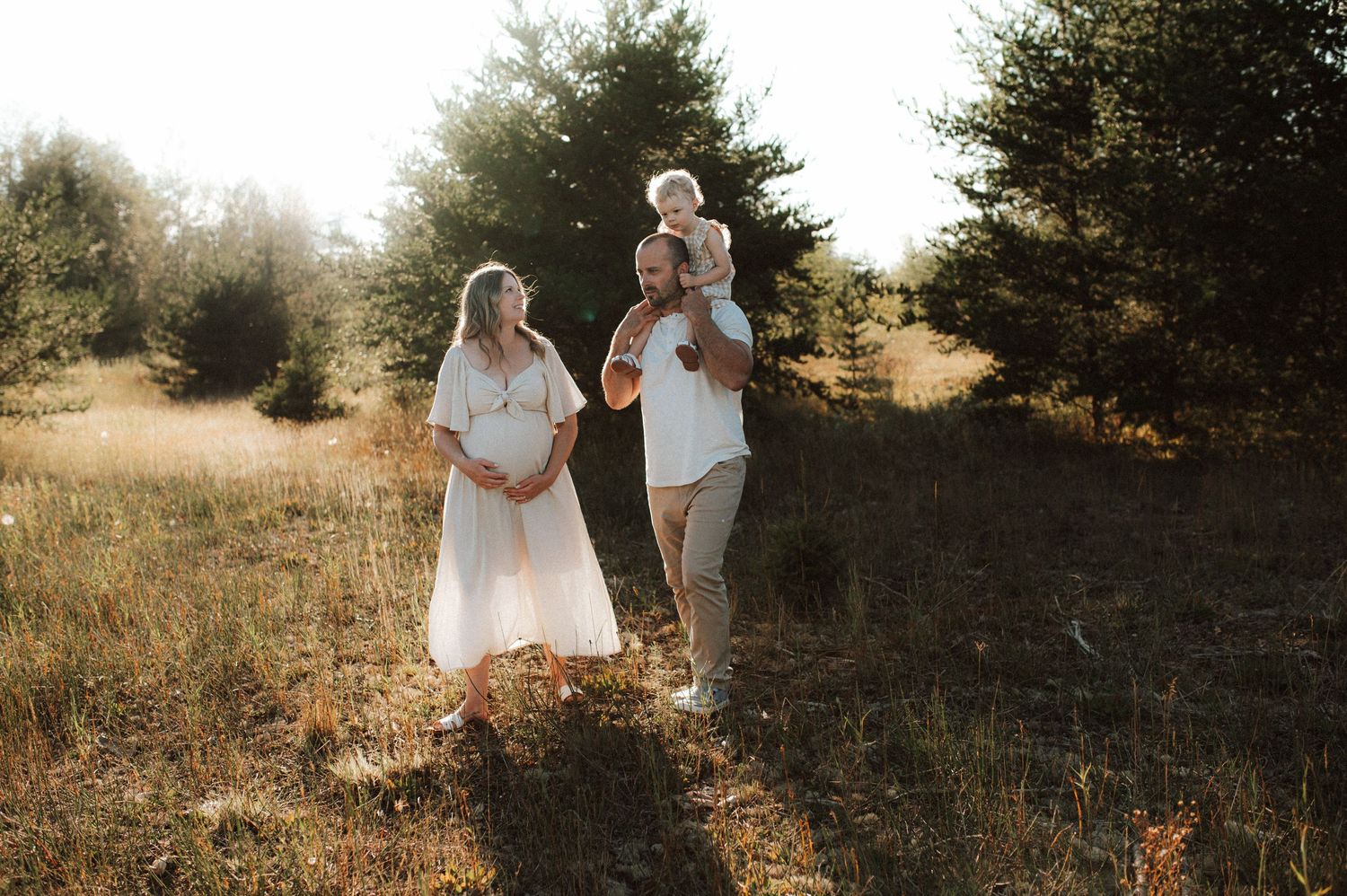 Couple stands together in a sunlit meadow at golden hour while wearing white and gray casual attire.