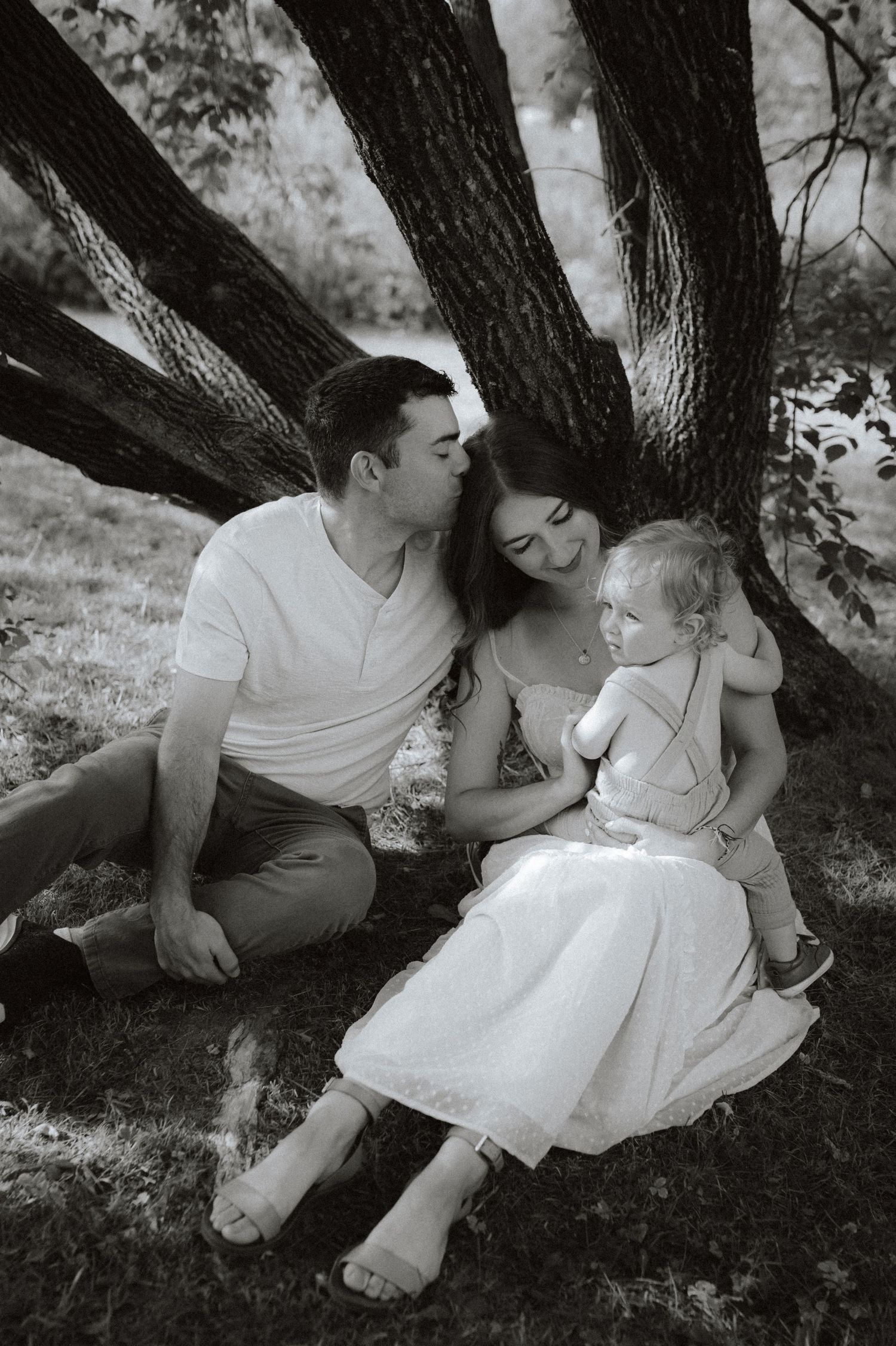 A family sits together in black and white under a large tree trunk, sharing an intimate moment in nature.