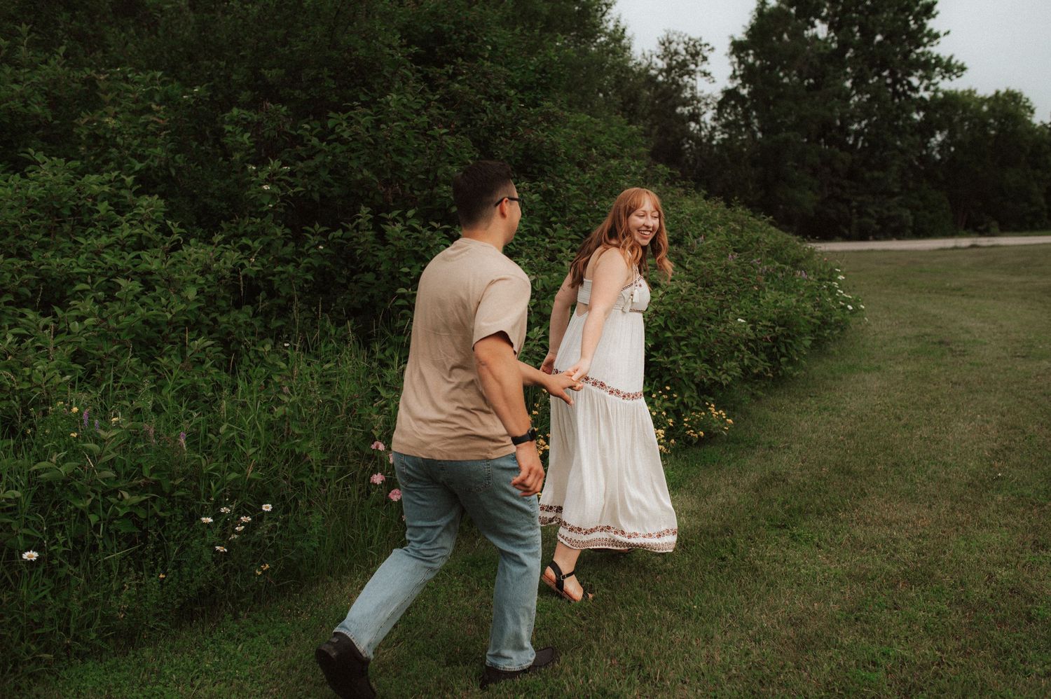 Two people walking together hand in hand along a lush green path surrounded by bushes and trees on a sunny day.