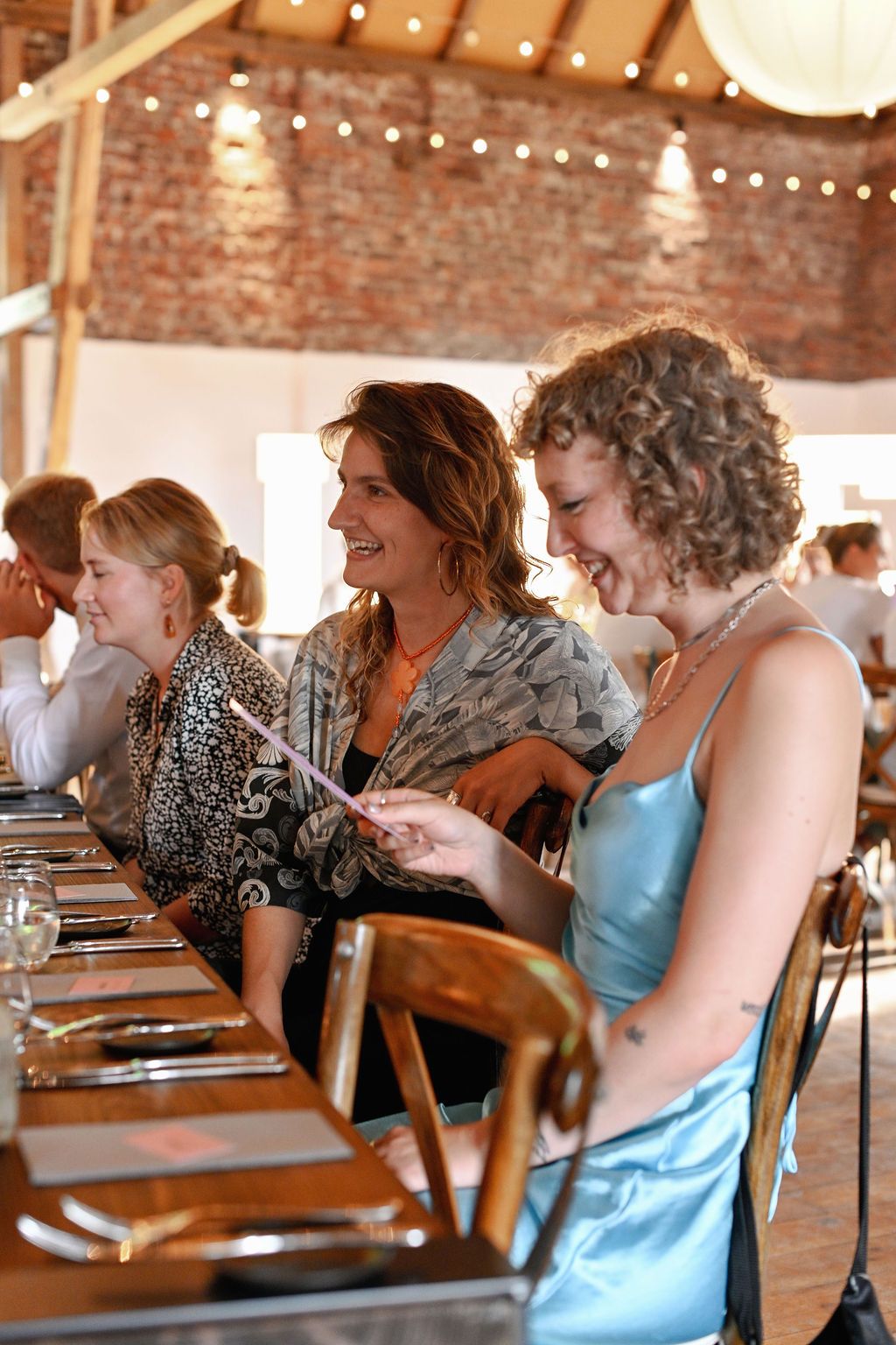 A group of people sit in wooden chairs at a rustic venue with exposed brick walls and string lights overhead.