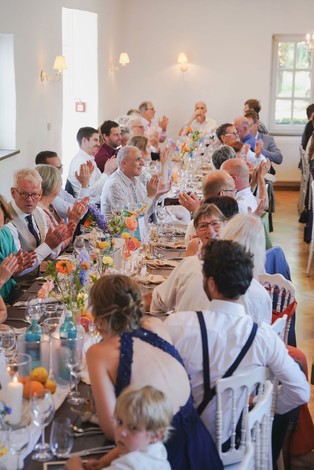 Wedding guests seated at long tables decorated with colorful flowers and glassware applaud during a reception celebration.