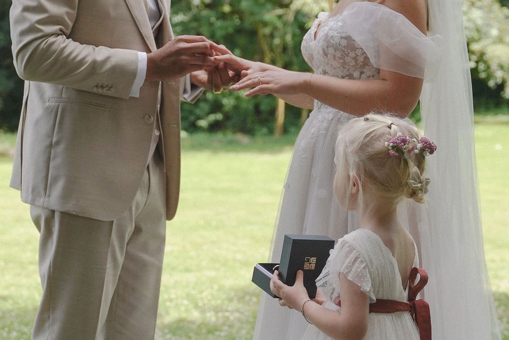 Couple exchanges rings during outdoor wedding ceremony while guests look on in a lush garden setting.