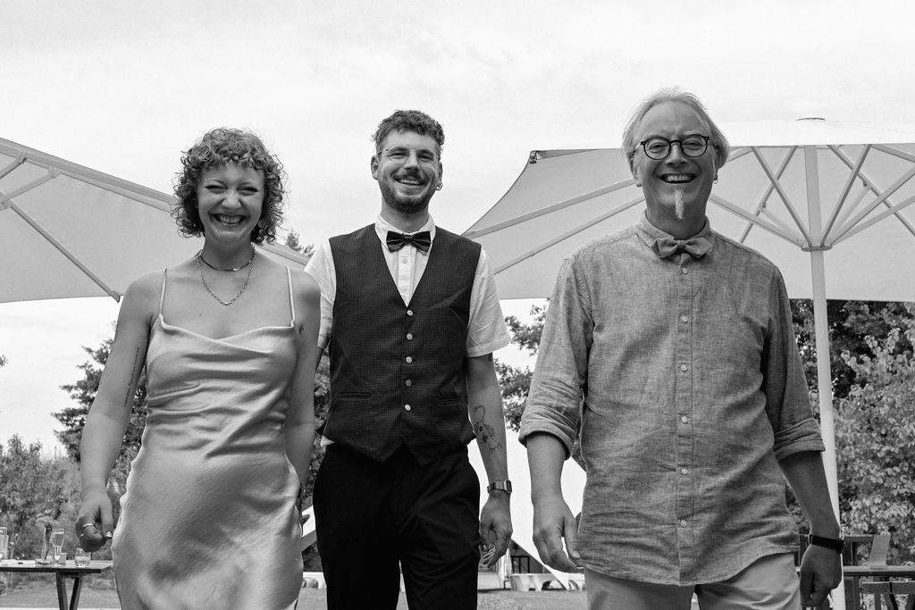 Three people stand together outdoors under white umbrellas at a formal event in black and white photography.