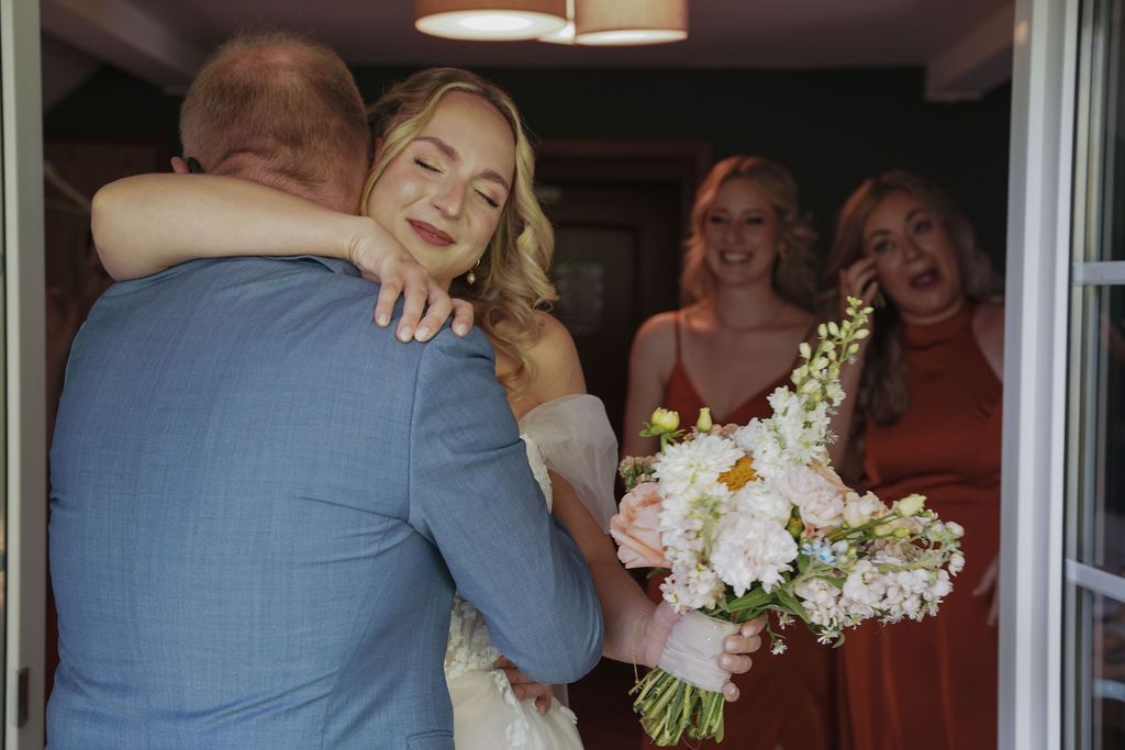 Wedding party members share embraces and smiles while wearing light blue and white formal attire.