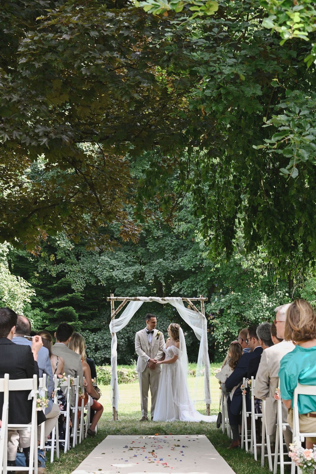 A wedding ceremony takes place under a white arch in a lush garden setting with white chairs and scattered rose petals.