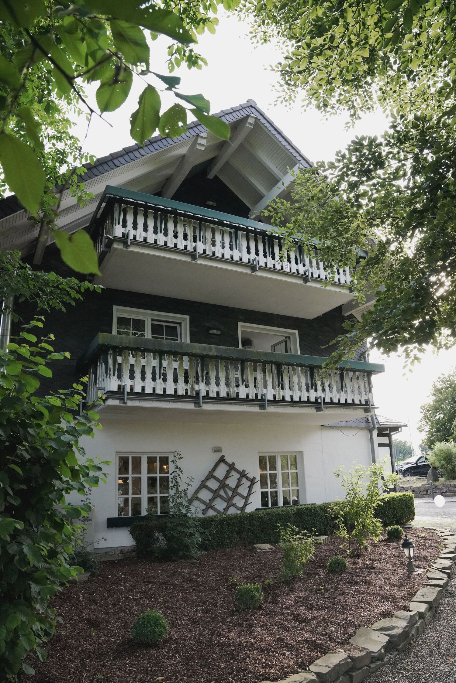 White mountain chalet-style house with decorative trim and windows nestled among green trees on a landscaped slope.