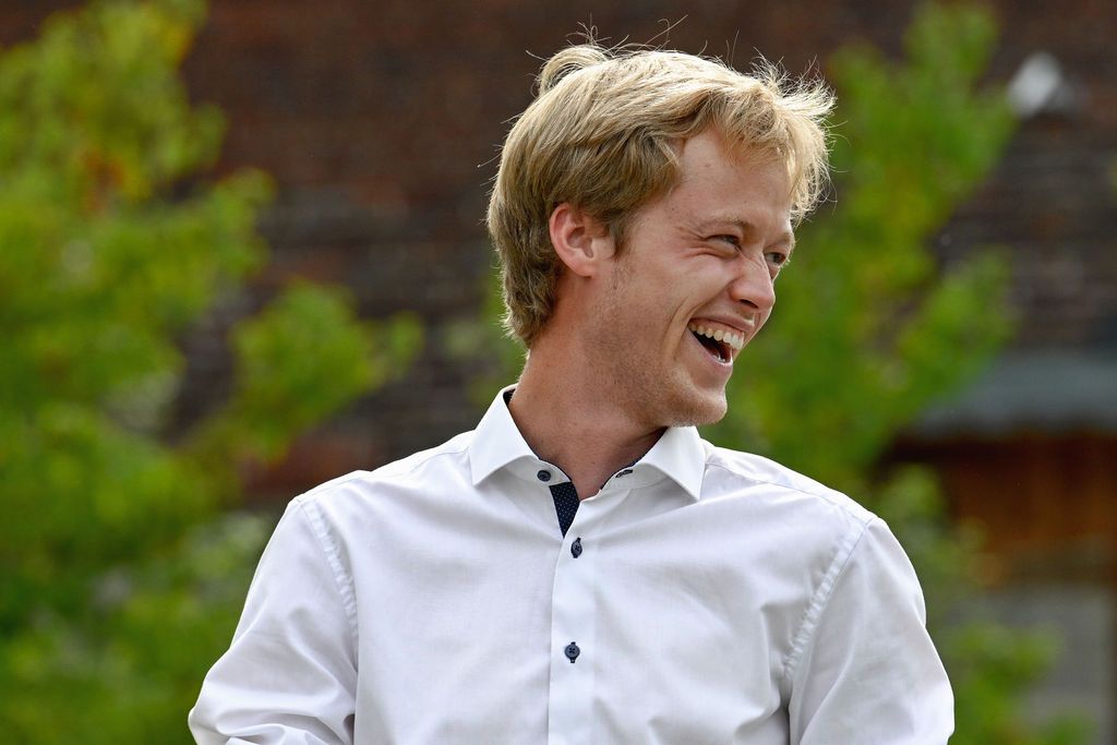 A person in a white button-down shirt smiles while sitting outdoors against a green natural background.