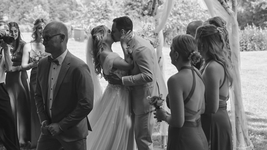 Black and white photograph of a wedding ceremony outdoors with wedding party members and guests gathered in a garden setting.