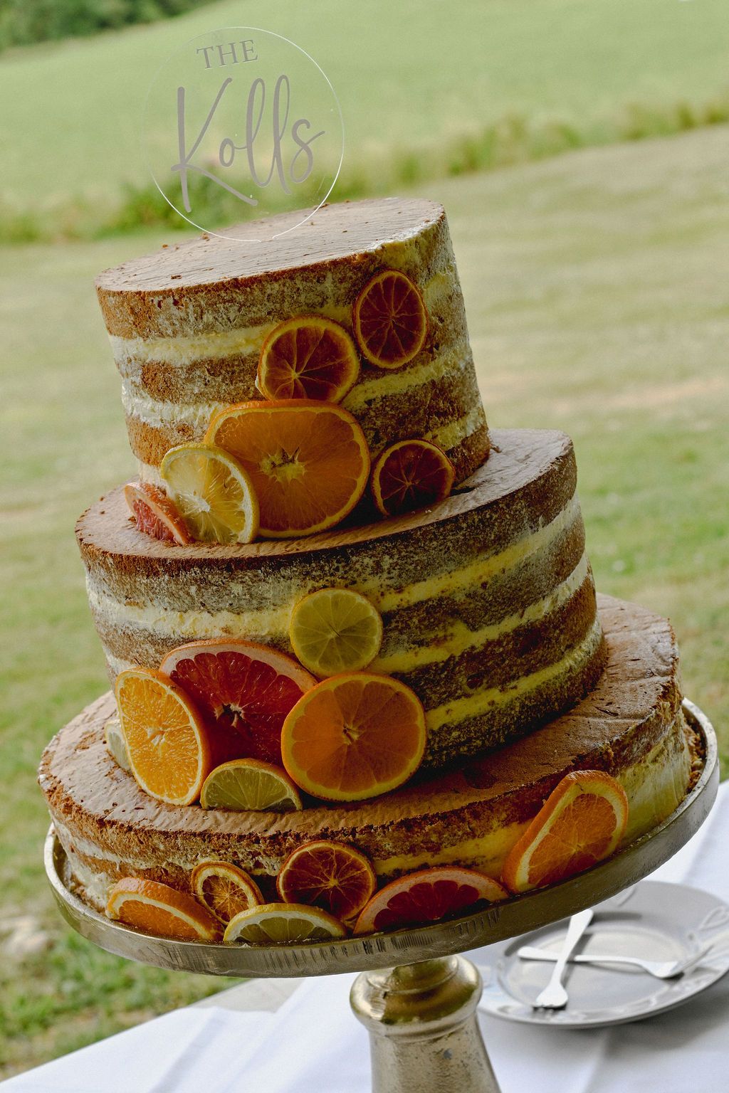 A rustic naked cake decorated with fresh citrus slices on a vintage cake stand in an outdoor setting.