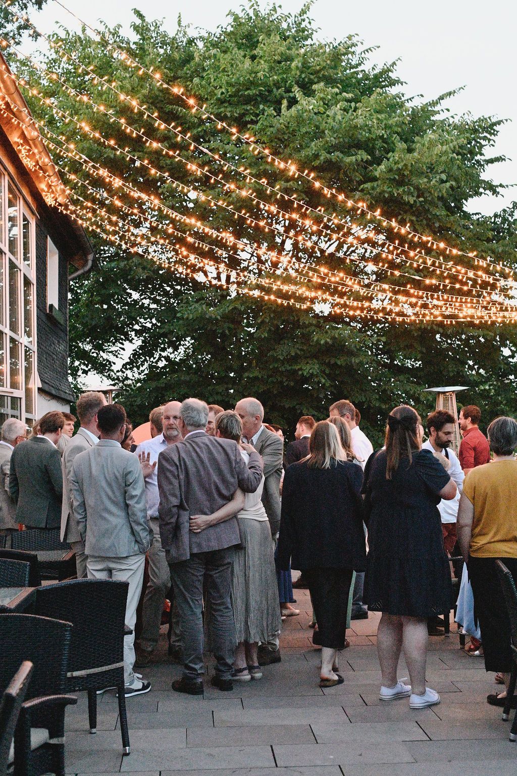 Guests mingle under twinkling string lights at an outdoor evening event with trees in the background.