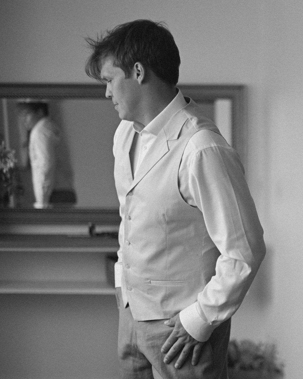 A black and white photograph shows a child in a white dress shirt and vest standing against a plain wall.