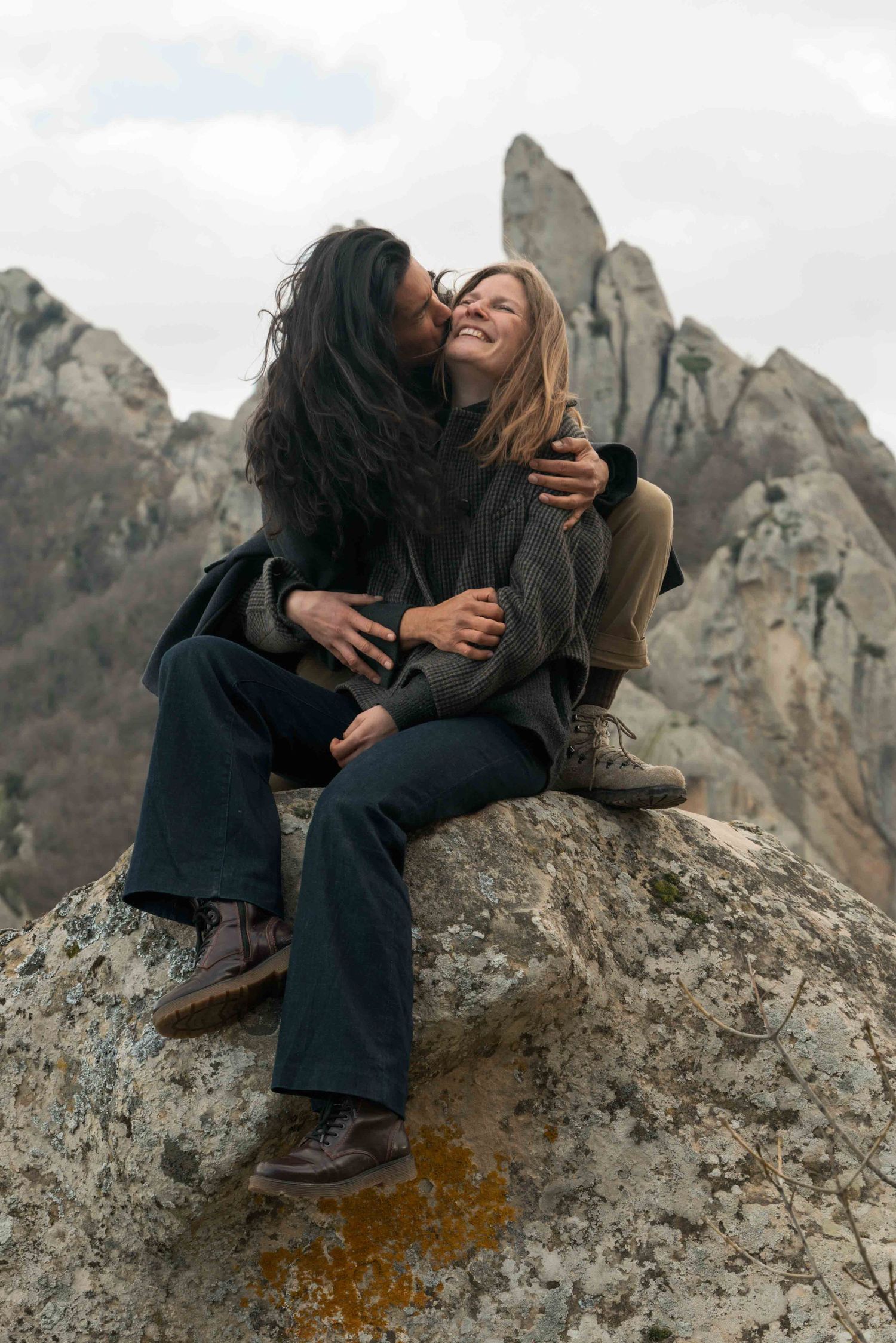 Two women embrace and laugh while sitting on a rocky mountain summit with jagged peaks in the background.