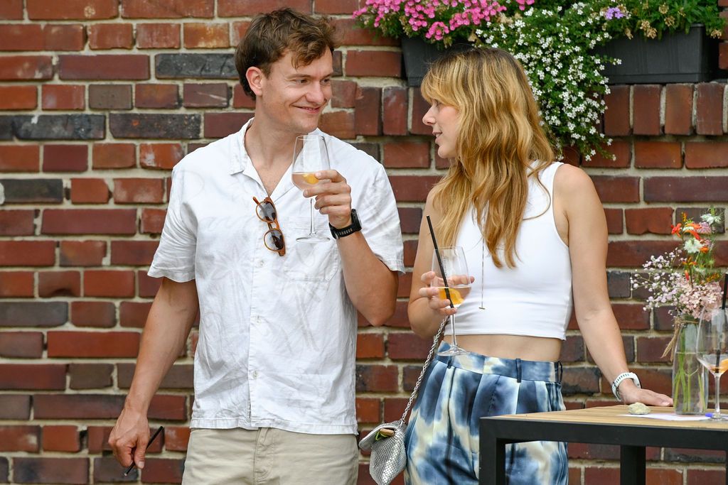 Two people enjoy drinks while standing by a brick wall with flowering plants on a sunny day.