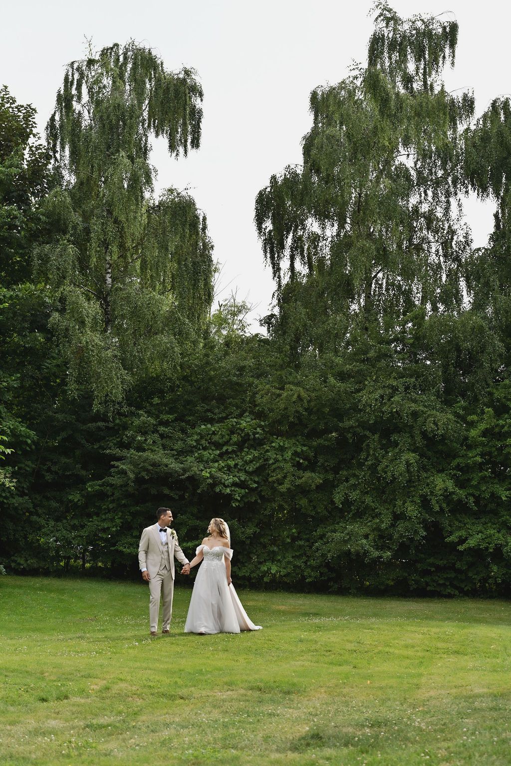 A couple in wedding attire stands together on a green lawn with weeping willow trees in the background.