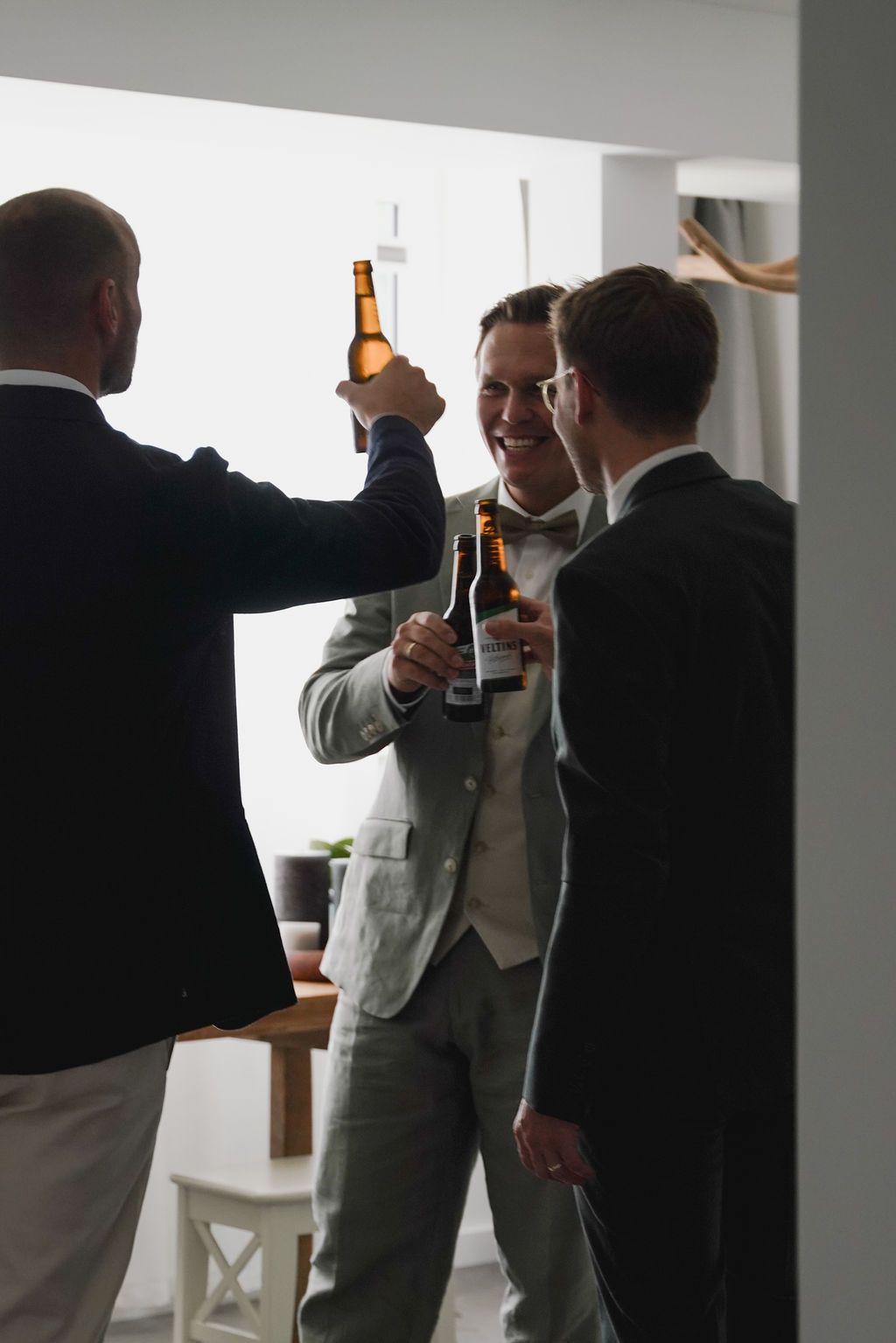 Three people in formal attire raise beer bottles in a celebratory toast against a bright window background.
