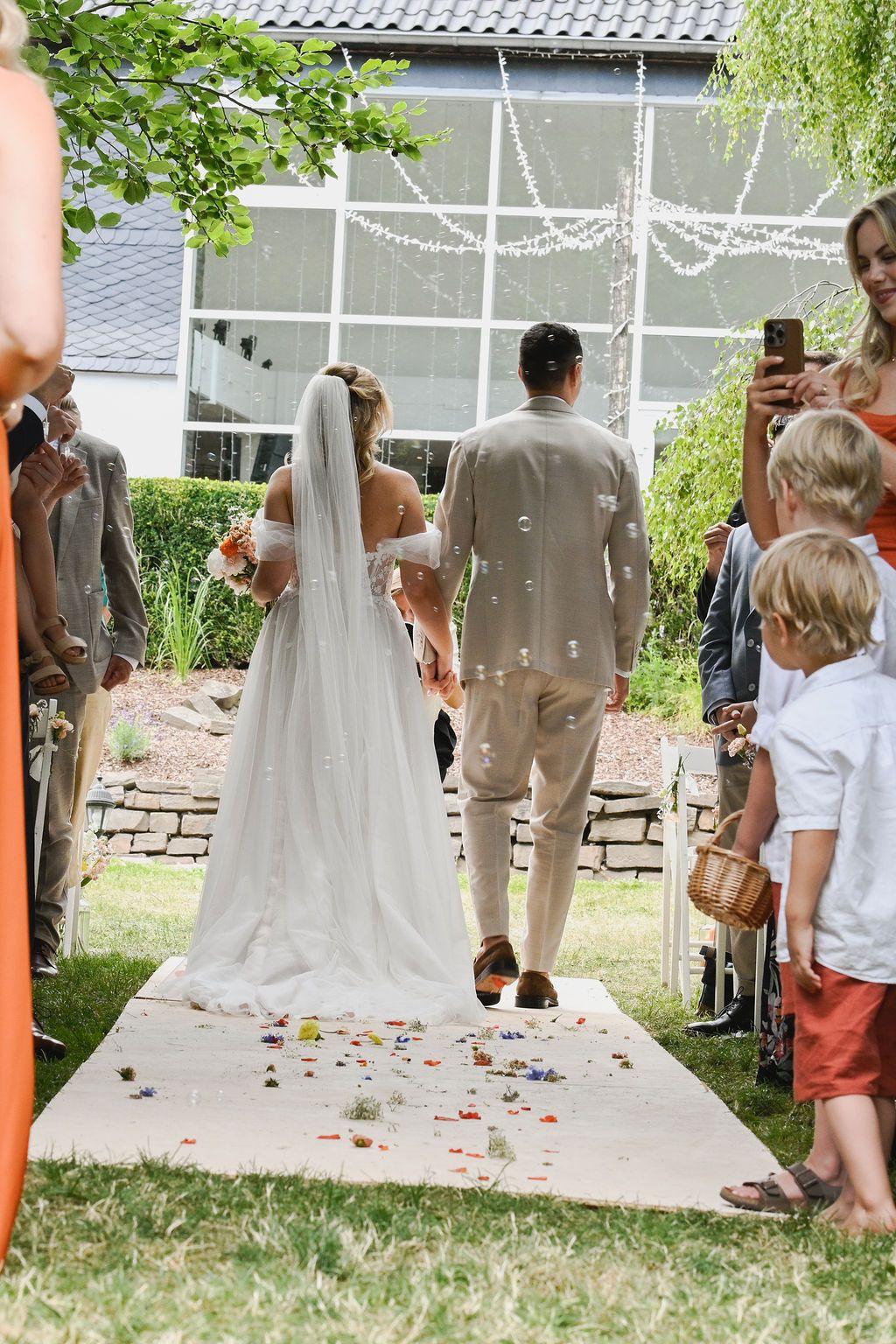 Wedding guests gather outdoors under orange poles with white fabric draping overhead during a garden ceremony.