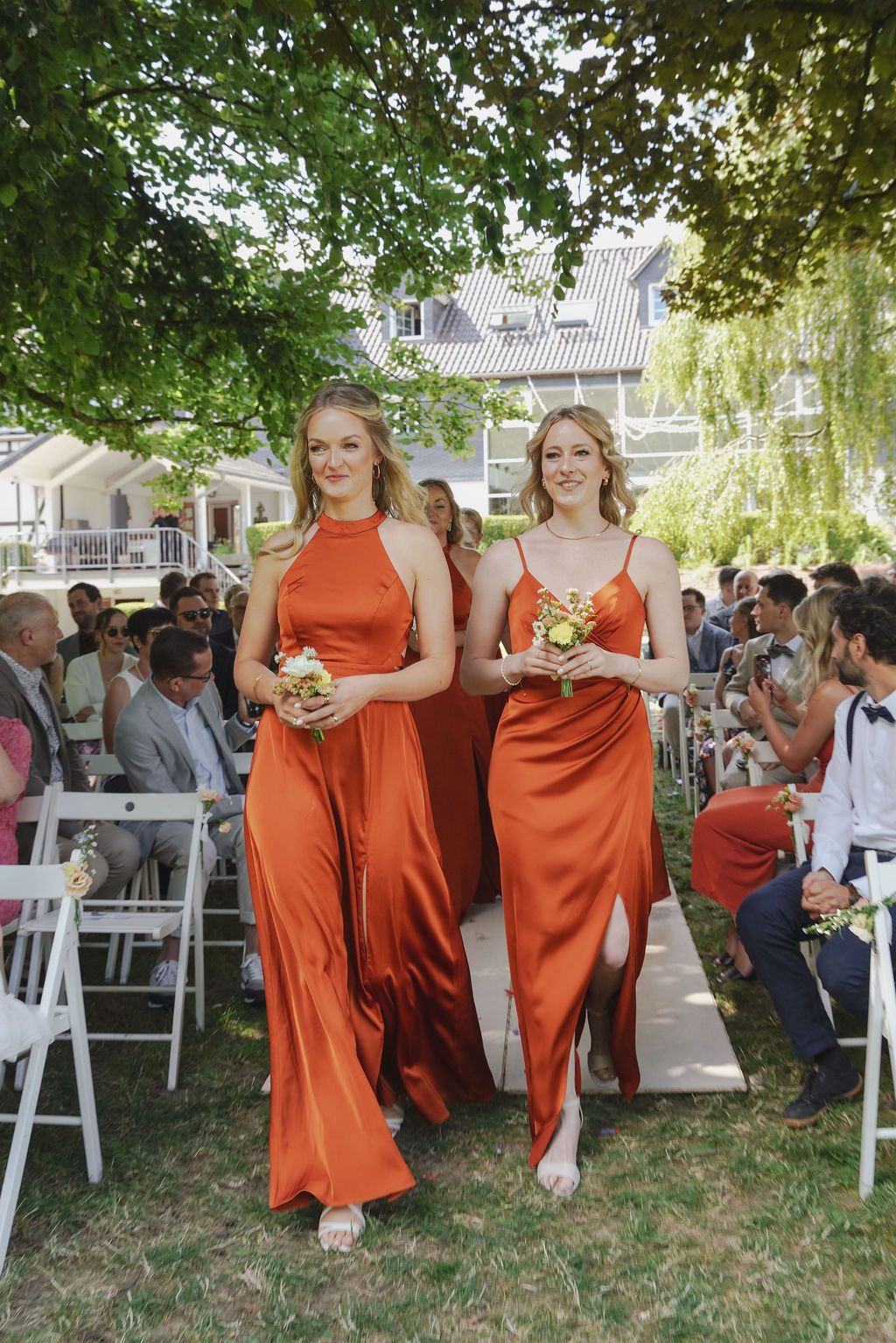 Two bridesmaids in flowing burnt orange dresses walk down an outdoor wedding aisle carrying bouquets.