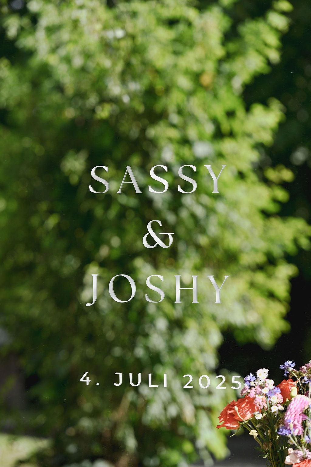 Wedding invitation design with text 'Sassy & Joshy' and date against blurred green foliage with flowers.