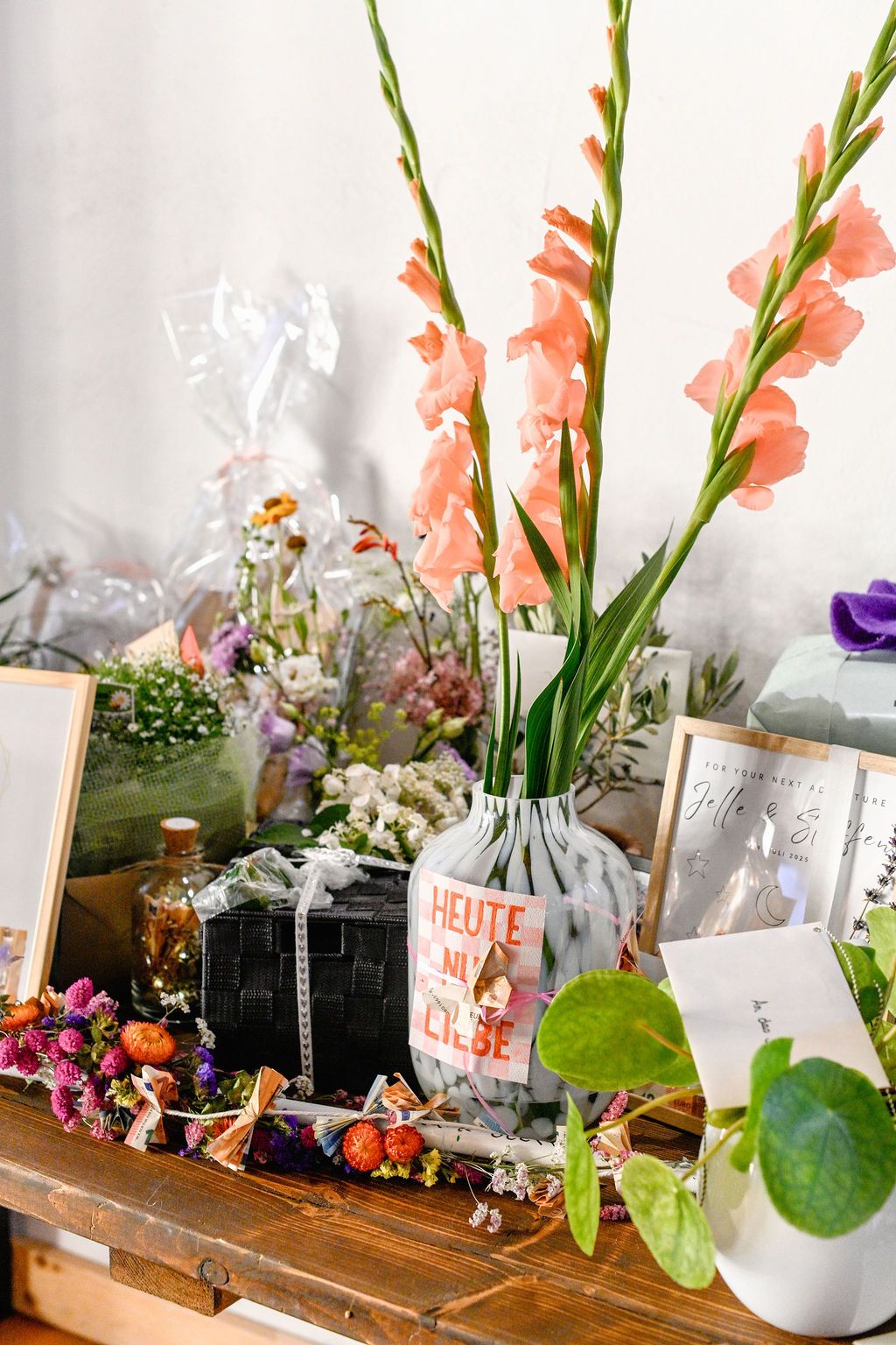 Pink gladiolus flowers in a vase alongside decorative items and greenery on a wooden surface with vintage styling.