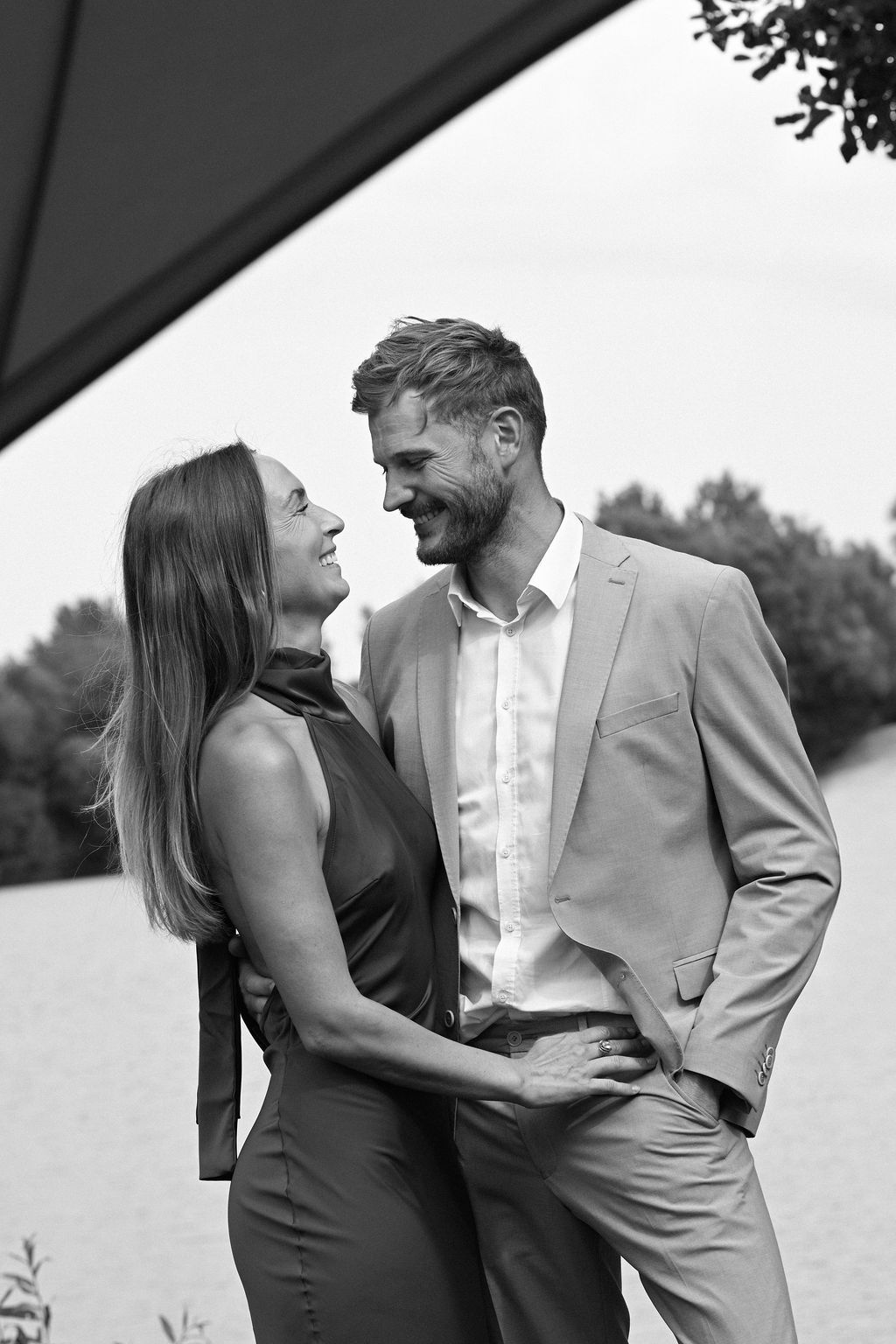 A romantic couple in formal attire shares intimate moments by a waterfront in black and white photography.