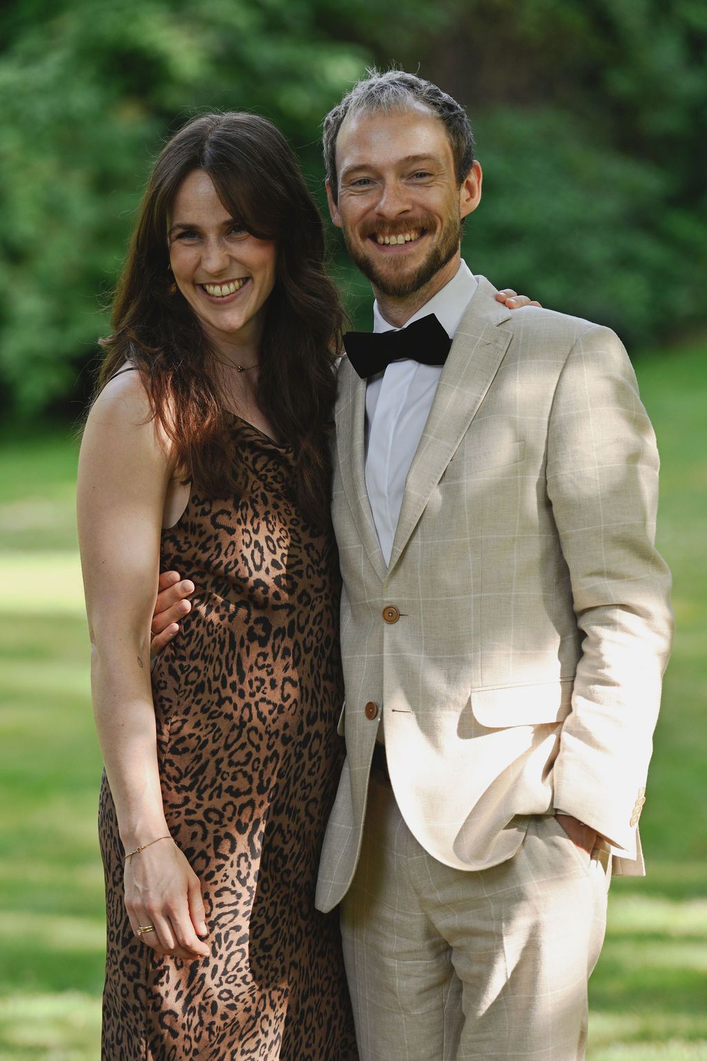 A couple stands together in a park wearing contrasting leopard print dress and light colored suit.