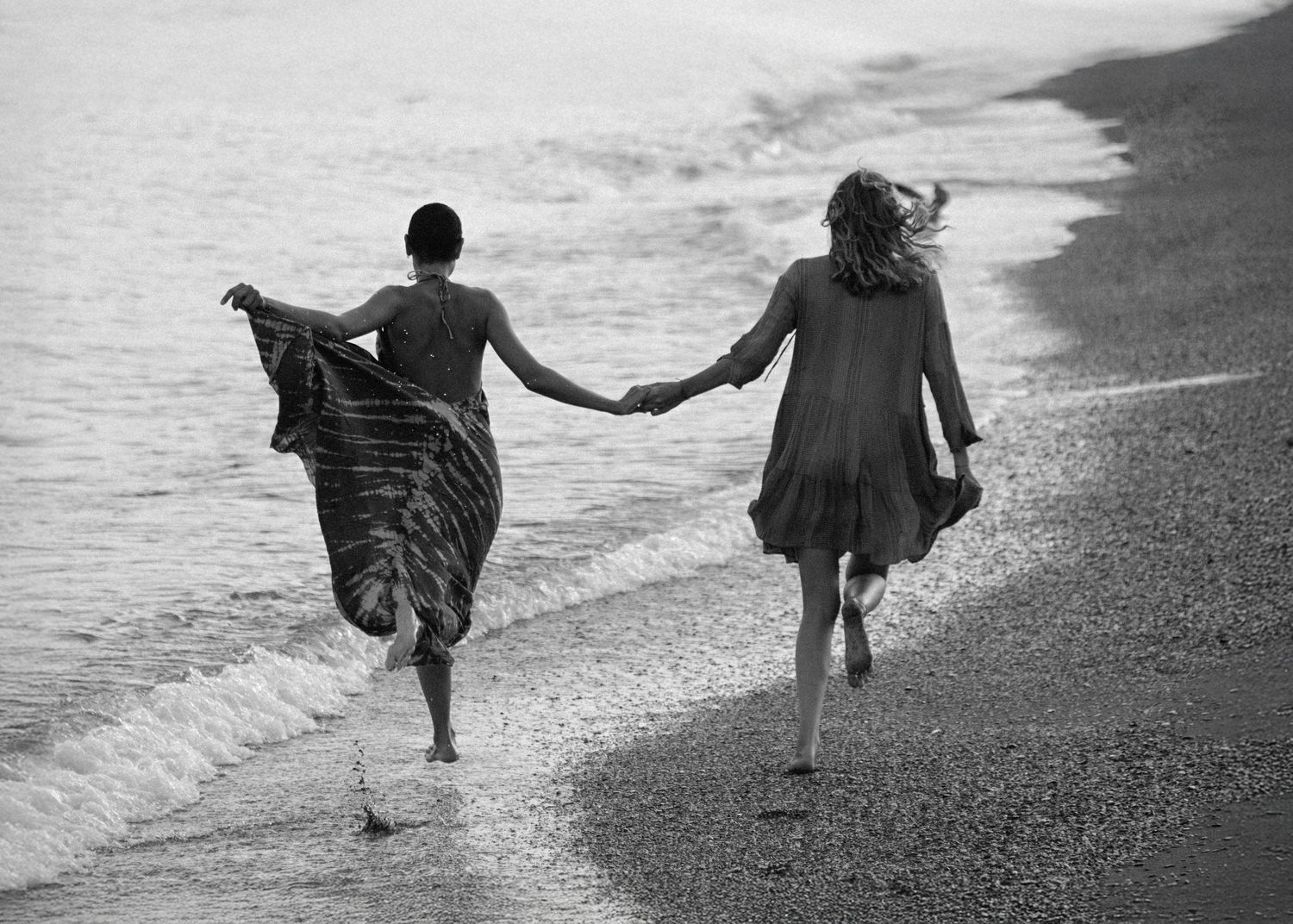 Two figures holding hands while walking along a beach shoreline in a black and white photograph.