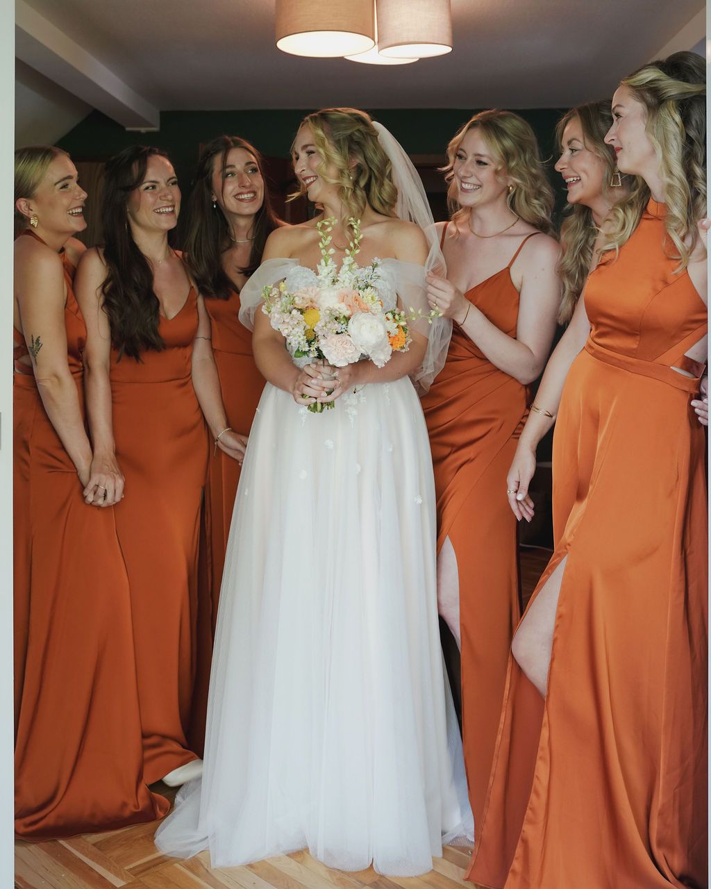 A bride in a white gown stands with bridesmaids wearing matching burnt orange dresses while holding a white floral bouquet.