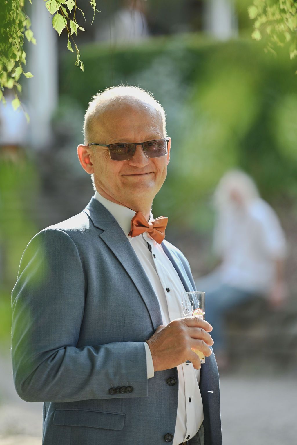A guest at an outdoor event wearing a grey suit with a bow tie and sunglasses holds a champagne glass.