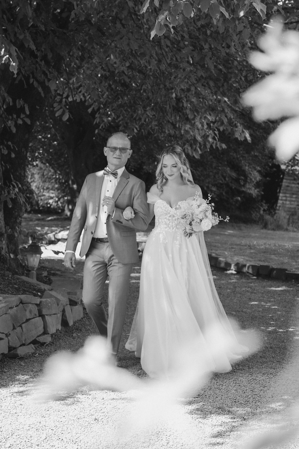 A black and white wedding photo showing a couple walking together on a garden path surrounded by trees and foliage.