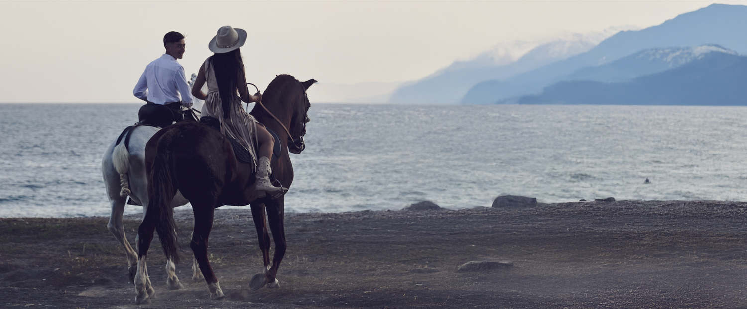 Two riders on horseback silhouetted against a misty coastal mountain landscape at dusk.