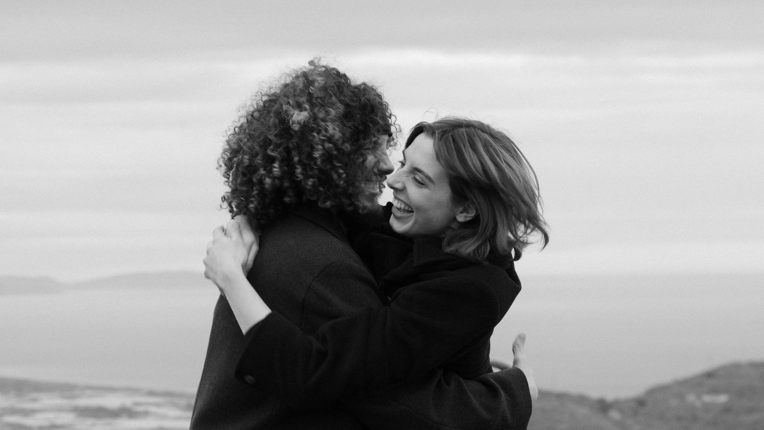 Two people sharing a warm embrace outdoors, smiling joyfully in a black and white photo with a scenic backdrop.