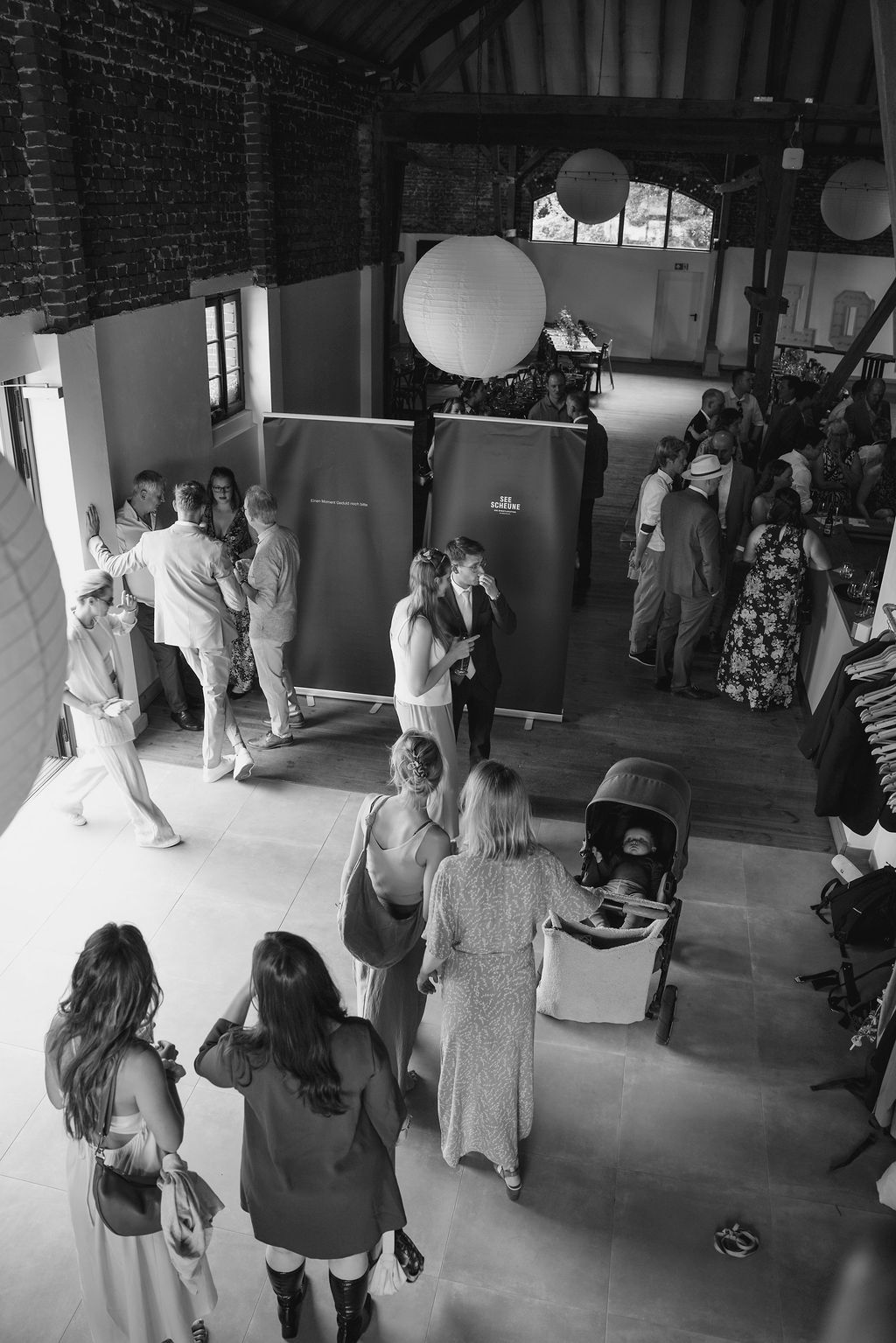 Wedding guests mingle and dance in a rustic indoor venue decorated with white paper lanterns.