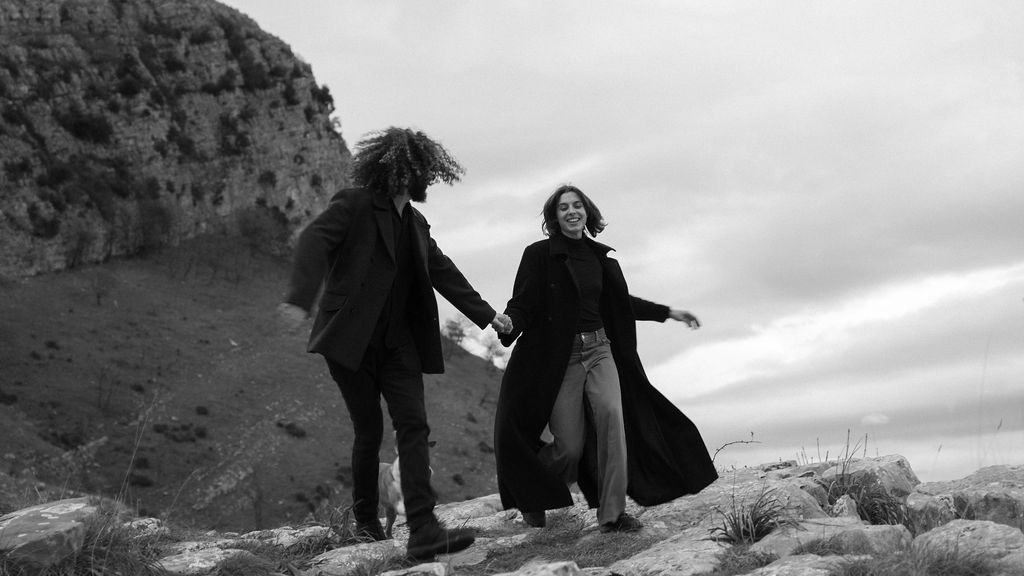 Two people in dark clothing hold hands while dancing against a dramatic mountain landscape in black and white.