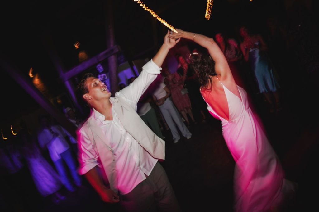 Energetic dancers with raised arms move to the music under purple and pink lighting at a nightclub party.