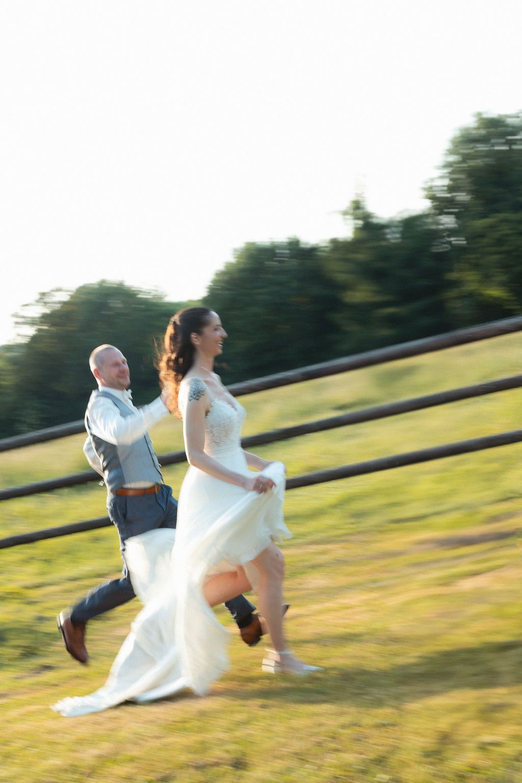 Bride and groom running joyfully through a sunlit field in wedding attire, motion blur capturing their happiness.