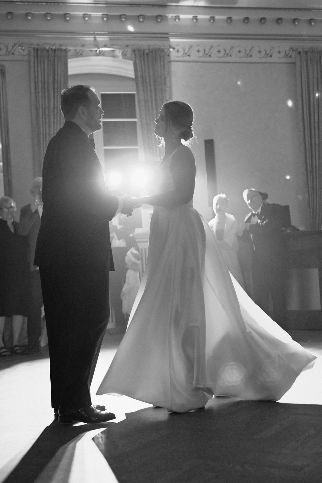 Black and white photo of bride and groom sharing first dance, illuminated by spotlight at elegant wedding reception.