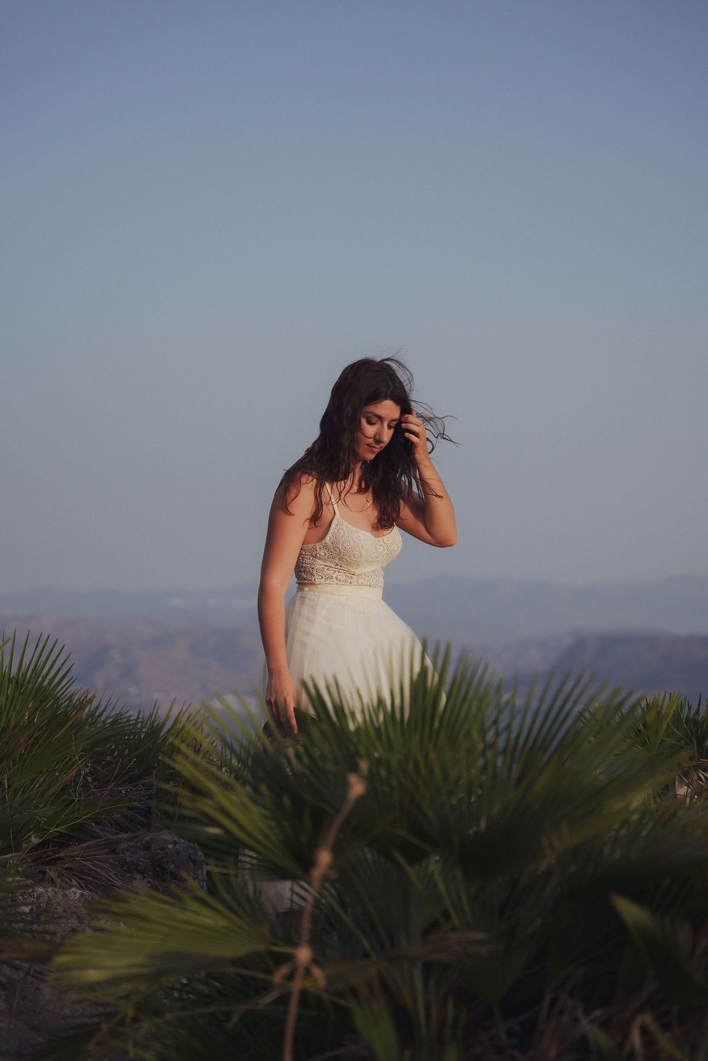 A bride in a white gown stands among palm plants at golden hour with mountains in the background.