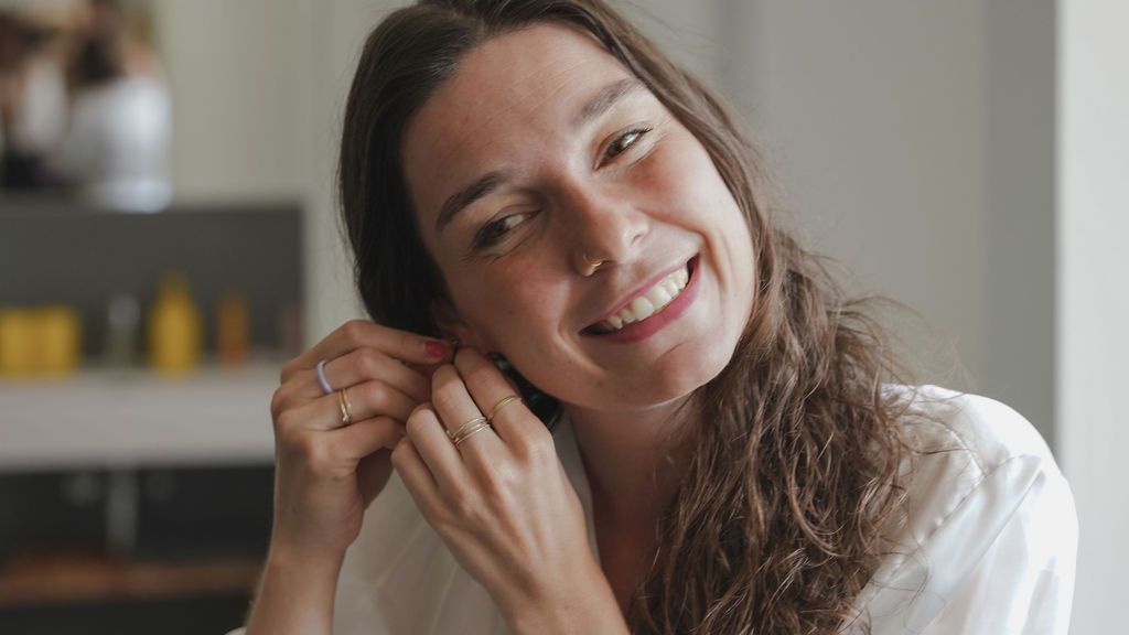 A person with wavy brown hair poses playfully in a white shirt against a light background.