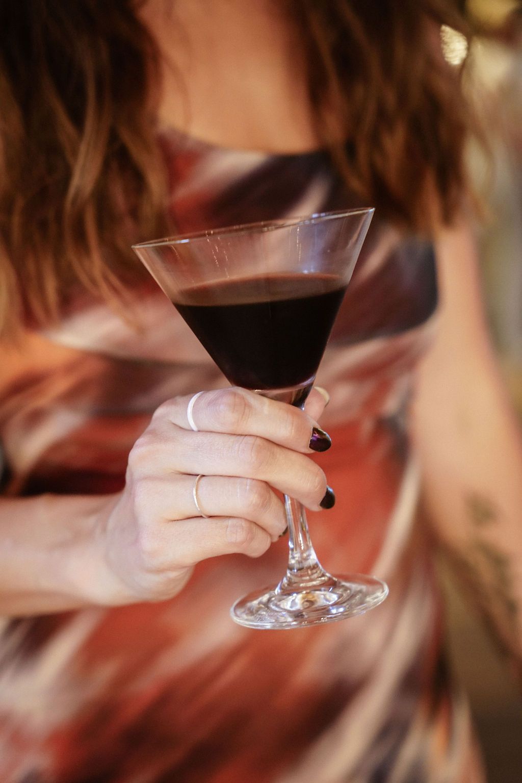 A hand with black nail polish holds a stemmed glass of red wine against a warm blurred background.