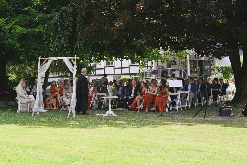 Outdoor wedding reception setup with white chairs and tables under shady trees on a grassy lawn.