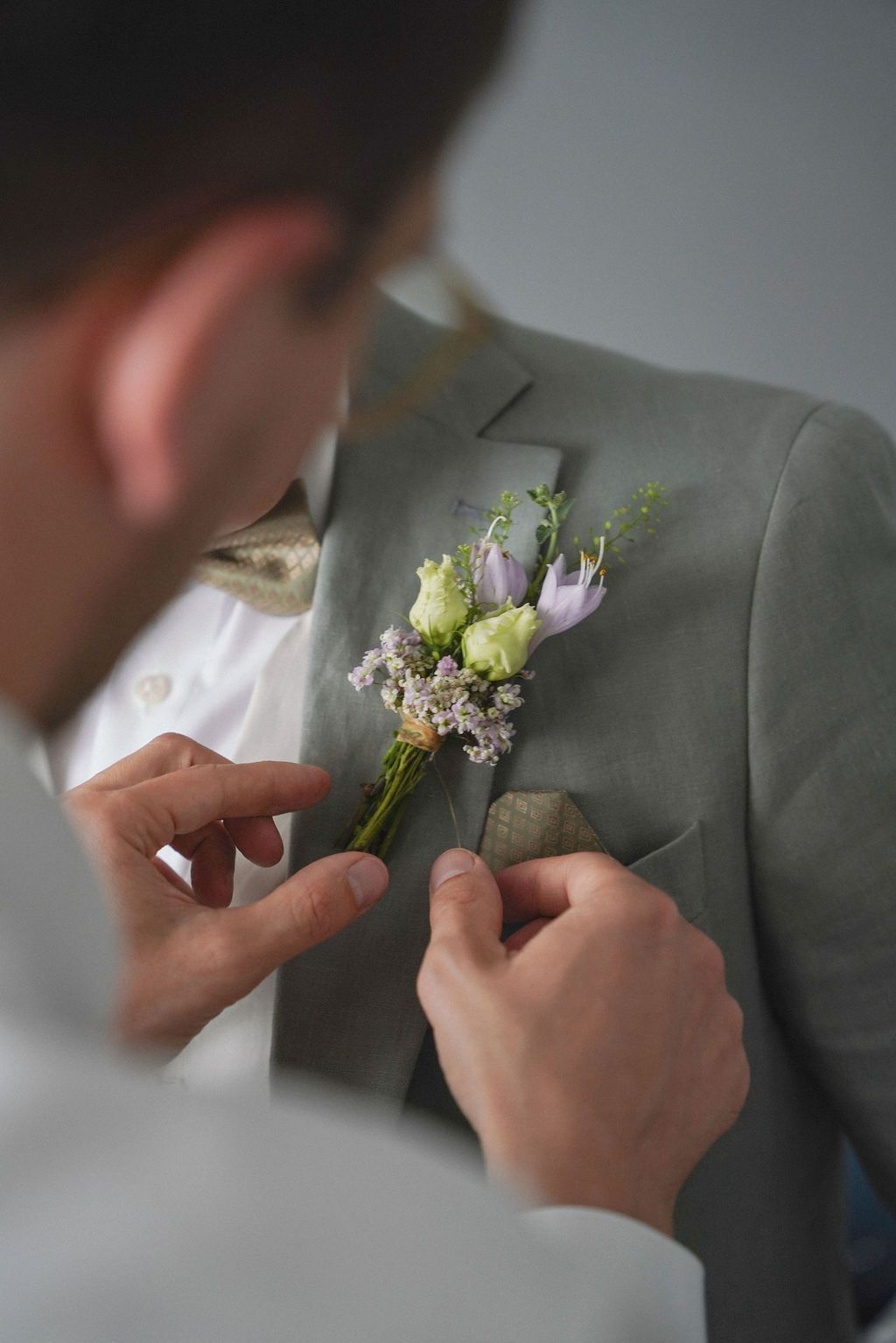 Groomsman pinning a floral boutonniere with green and purple flowers onto a gray suit lapel at a wedding.