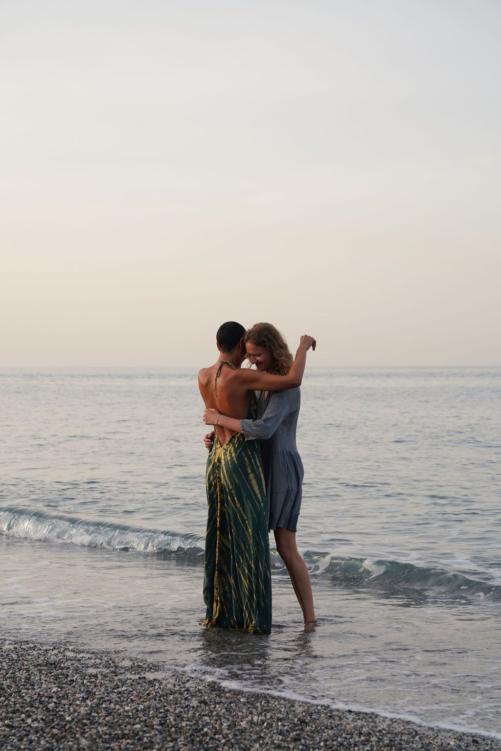 Romantic couple embracing on a pebble beach at dusk, standing in shallow ocean waves with calm sea in background.