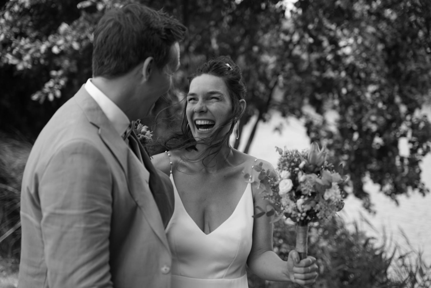 Black and white photo of a laughing bride holding flowers beside her groom at an outdoor wedding ceremony.