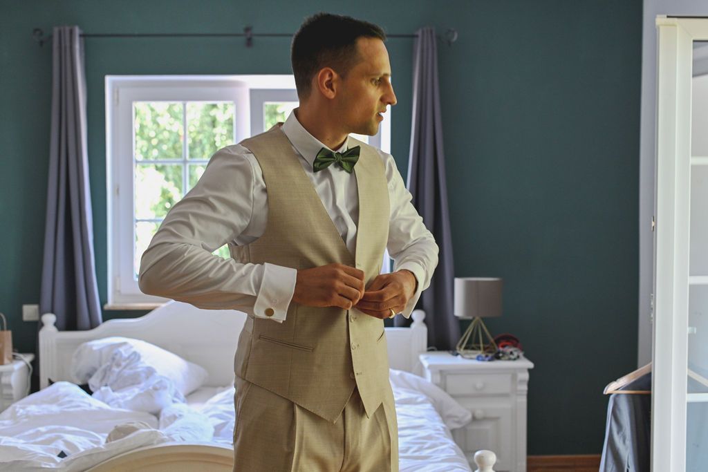 Someone in a light beige suit adjusts their jacket while standing in a bedroom with teal walls and natural lighting.