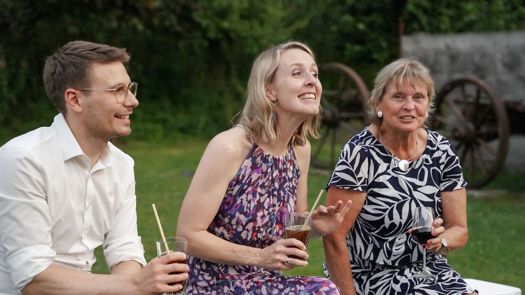Three people sitting outdoors share a laugh while holding drinks at a casual summer gathering.